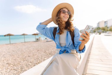 Şapka takan ve sırt çantası taşıyan bir gezgin kumsalın yanındaki bankta oturmuş, elinde telefon tutuyordu. Telefonu olan genç bir kadın gün batımında deniz manzarasının tadını çıkarıyor. Turizm, eğlence ve doğa için kavramsal fikirler.
