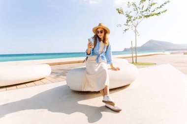 Şapka takan ve sırt çantası taşıyan bir gezgin kumsalın yanındaki bankta oturmuş, elinde telefon tutuyordu. Telefonu olan genç bir kadın gün batımında deniz manzarasının tadını çıkarıyor. Turizm, eğlence ve doğa için kavramsal fikirler.