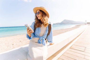 Şapka takan ve sırt çantası taşıyan bir gezgin kumsalın yanındaki bankta oturmuş, elinde telefon tutuyordu. Telefonu olan genç bir kadın gün batımında deniz manzarasının tadını çıkarıyor. Turizm, eğlence ve doğa için kavramsal fikirler.