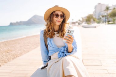 Şapka takan ve sırt çantası taşıyan bir gezgin kumsalın yanındaki bankta oturmuş, elinde telefon tutuyordu. Telefonu olan genç bir kadın gün batımında deniz manzarasının tadını çıkarıyor. Turizm, eğlence ve doğa için kavramsal fikirler.