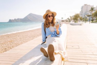 Şapka takan ve sırt çantası taşıyan bir gezgin kumsalın yanındaki bankta oturmuş, elinde telefon tutuyordu. Telefonu olan genç bir kadın gün batımında deniz manzarasının tadını çıkarıyor. Turizm, eğlence ve doğa için kavramsal fikirler.