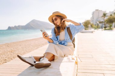 Şapka takan ve sırt çantası taşıyan bir gezgin kumsalın yanındaki bankta oturmuş, elinde telefon tutuyordu. Telefonu olan genç bir kadın gün batımında deniz manzarasının tadını çıkarıyor. Turizm, eğlence ve doğa için kavramsal fikirler.