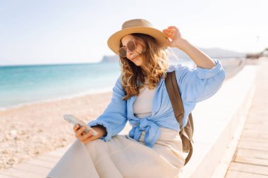 Şapka takan ve sırt çantası taşıyan bir gezgin kumsalın yanındaki bankta oturmuş, elinde telefon tutuyordu. Telefonu olan genç bir kadın gün batımında deniz manzarasının tadını çıkarıyor. Turizm, eğlence ve doğa için kavramsal fikirler.