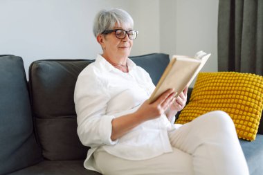 Gri saçlı yetişkin bir kadın rahat bir odada kanepede otururken kitap okur. Emekli bir kadın oturma odasında kitap okuyarak rahatlıyor. Rahatlama, emeklilik ve okuma kavramları.