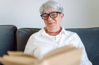 Gri saçlı yetişkin bir kadın rahat bir odada kanepede otururken kitap okur. Emekli bir kadın oturma odasında kitap okuyarak rahatlıyor. Rahatlama, emeklilik ve okuma kavramları.