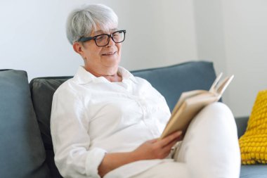 Gri saçlı yetişkin bir kadın rahat bir odada kanepede otururken kitap okur. Emekli bir kadın oturma odasında kitap okuyarak rahatlıyor. Rahatlama, emeklilik ve okuma kavramları.