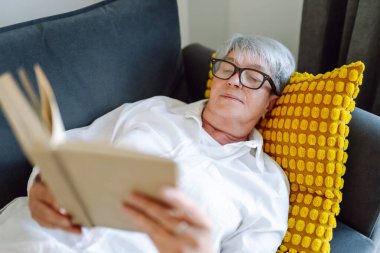 Gri saçlı yetişkin bir kadın rahat bir odada kanepede otururken kitap okur. Emekli bir kadın oturma odasında kitap okuyarak rahatlıyor. Rahatlama, emeklilik ve okuma kavramları.