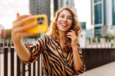 Elinde telefonla gülümseyen bir kadın, metropolün arka planına karşı blog yazıyor ya da video konferans yapıyor. Turistler elinde bir telefonla günbatımı gezintisini severler. Selfie zamanı! Turizm ve blog kavramları.