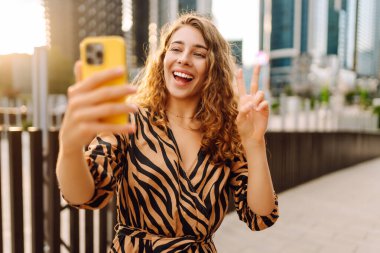 Elinde telefonla gülümseyen bir kadın, metropolün arka planına karşı blog yazıyor ya da video konferans yapıyor. Turistler elinde bir telefonla günbatımı gezintisini severler. Selfie zamanı! Turizm ve blog kavramları.