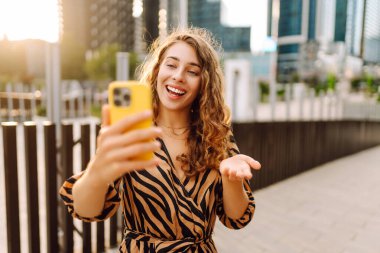 Elinde telefonla gülümseyen bir kadın, metropolün arka planına karşı blog yazıyor ya da video konferans yapıyor. Turistler elinde bir telefonla günbatımı gezintisini severler. Selfie zamanı! Turizm ve blog kavramları.