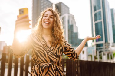 Elinde telefonla gülümseyen bir kadın, metropolün arka planına karşı blog yazıyor ya da video konferans yapıyor. Turistler elinde bir telefonla günbatımı gezintisini severler. Selfie zamanı! Turizm ve blog kavramları.