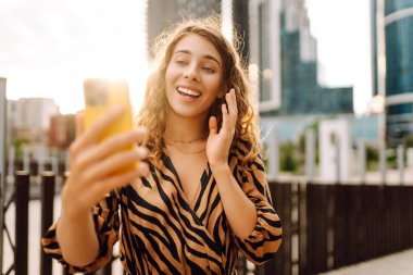 Elinde telefonla gülümseyen bir kadın, metropolün arka planına karşı blog yazıyor ya da video konferans yapıyor. Turistler elinde bir telefonla günbatımı gezintisini severler. Selfie zamanı! Turizm ve blog kavramları.