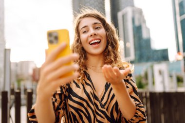 Elinde telefonla gülümseyen bir kadın, metropolün arka planına karşı blog yazıyor ya da video konferans yapıyor. Turistler elinde bir telefonla günbatımı gezintisini severler. Selfie zamanı! Turizm ve blog kavramları.