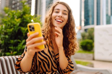 Elinde telefonla gülümseyen bir kadın, metropolün arka planına karşı blog yazıyor ya da video konferans yapıyor. Turistler elinde bir telefonla günbatımı gezintisini severler. Selfie zamanı! Turizm ve blog kavramları.
