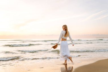 Genç bir kadın gün doğumunda plaj boyunca yürür. Şapkalı neşeli bir kadın açık deniz manzarasını seviyor. Doğa kavramları, rahatlama ve hafta sonları..