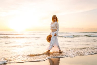 Genç bir kadın gün doğumunda plaj boyunca yürür. Şapkalı neşeli bir kadın açık deniz manzarasını seviyor. Doğa kavramları, rahatlama ve hafta sonları..