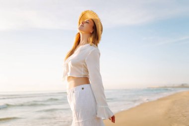 Genç bir kadın gün doğumunda plaj boyunca yürür. Şapkalı neşeli bir kadın açık deniz manzarasını seviyor. Doğa kavramları, rahatlama ve hafta sonları..