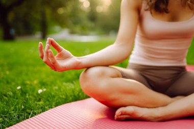 Atletik giysili barışçıl bir kadın gün batımında bir parkta yoga minderi üzerinde meditasyon yapıyor. Genç bir kadın yoga yapıyor ve açık havada güneşli bir günün tadını çıkarıyor. Rahatlama, meditasyon ve hafta sonları için kavramlar.