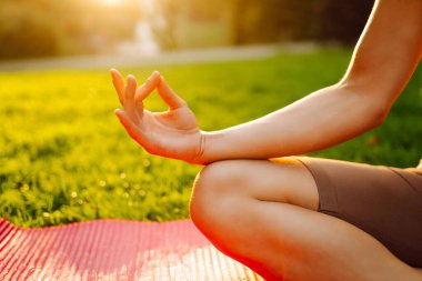Atletik giysili barışçıl bir kadın gün batımında bir parkta yoga minderi üzerinde meditasyon yapıyor. Genç bir kadın yoga yapıyor ve açık havada güneşli bir günün tadını çıkarıyor. Rahatlama, meditasyon ve hafta sonları için kavramlar.