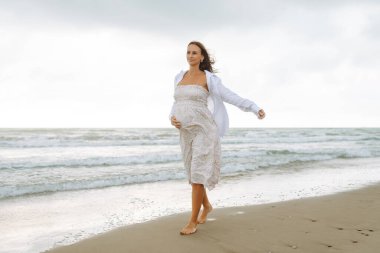 Elbiseli hamile bir kadın bulutlu bir günde deniz kenarında yürümekten hoşlanır. Çocuk bekleyen güzel bir kadın kendini özgür hisseder ve dışarıda dinlenir. Doğa konsepti, yürü.