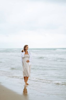 Elbiseli hamile bir kadın bulutlu bir günde deniz kenarında yürümekten hoşlanır. Çocuk bekleyen güzel bir kadın kendini özgür hisseder ve dışarıda dinlenir. Doğa konsepti, yürü.