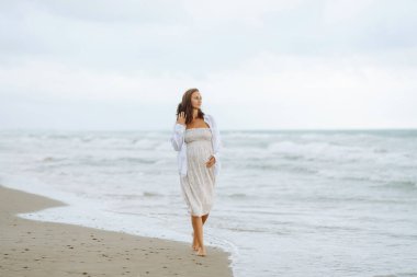 Elbiseli hamile bir kadın bulutlu bir günde deniz kenarında yürümekten hoşlanır. Çocuk bekleyen güzel bir kadın kendini özgür hisseder ve dışarıda dinlenir. Doğa konsepti, yürü.