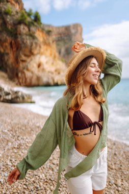 Hasır şapkalı neşeli bir kadın gün batımında çakıl taşlı bir sahilde deniz manzarasının tadını çıkarıyor. Mayolu genç bir kadın açık havada kendini özgür hissediyor. Rahatlama, doğa ve özgürlük kavramı.