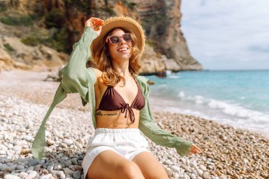 Hasır şapkalı neşeli bir kadın gün batımında çakıl taşlı bir sahilde deniz manzarasının tadını çıkarıyor. Mayolu genç bir kadın açık havada kendini özgür hissediyor. Rahatlama, doğa ve özgürlük kavramı.