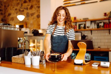 Tezgahın arkasında elinde bir fincan taze kahve tutan genç bir bayan barista var. Tecrübeli bir barista rahat bir kafede aromatik kahve hazırlar. İçkiler, konfor ve hazırlık konsepti.