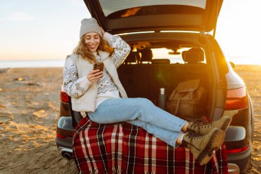 Şafak vakti gülümseyen bir kadın bir arabanın bagajında otururken telefonuyla blog yazıyor. Bir gezgin selfie çeker ve yolculuk sırasında deniz manzarasının tadını çıkarır. Bloglama ve ulaşım kavramları.