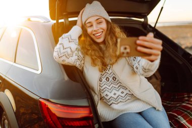 Şafak vakti gülümseyen bir kadın bir arabanın bagajında otururken telefonuyla blog yazıyor. Bir gezgin selfie çeker ve yolculuk sırasında deniz manzarasının tadını çıkarır. Bloglama ve ulaşım kavramları.