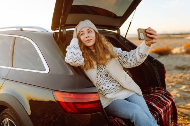 Şafak vakti gülümseyen bir kadın bir arabanın bagajında otururken telefonuyla blog yazıyor. Bir gezgin selfie çeker ve yolculuk sırasında deniz manzarasının tadını çıkarır. Bloglama ve ulaşım kavramları.