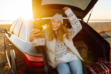 Şafak vakti gülümseyen bir kadın bir arabanın bagajında otururken telefonuyla blog yazıyor. Bir gezgin selfie çeker ve yolculuk sırasında deniz manzarasının tadını çıkarır. Bloglama ve ulaşım kavramları.