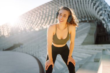 Spor kıyafetleri ve kablosuz kulaklıklar takan bir kadın dışarıda egzersiz yapıyor. Atletik bir kadın güneşli bir günde antrenmandan önce müzik dinler. Müzik ve spor konsepti. Yaşam biçimi.