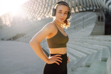 Spor kıyafetleri ve kablosuz kulaklıklar takan bir kadın dışarıda egzersiz yapıyor. Atletik bir kadın güneşli bir günde antrenmandan önce müzik dinler. Müzik ve spor konsepti. Yaşam biçimi.