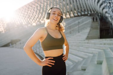 Spor kıyafetleri ve kablosuz kulaklıklar takan bir kadın dışarıda egzersiz yapıyor. Atletik bir kadın güneşli bir günde antrenmandan önce müzik dinler. Müzik ve spor konsepti. Yaşam biçimi.