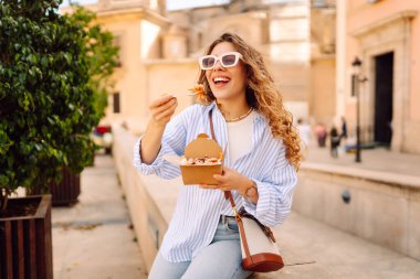 Neşeli bir kadın dışarıda aromatik makarna yer. Güneşli bir günde güzel bir turist bir paket kutusu tutar. Turizm, gıda ve eğlence kavramları.