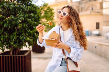 Neşeli bir kadın dışarıda aromatik makarna yer. Güneşli bir günde güzel bir turist bir paket kutusu tutar. Turizm, gıda ve eğlence kavramları.