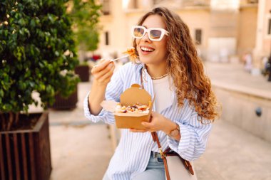 Neşeli bir kadın dışarıda aromatik makarna yer. Güneşli bir günde güzel bir turist bir paket kutusu tutar. Turizm, gıda ve eğlence kavramları.