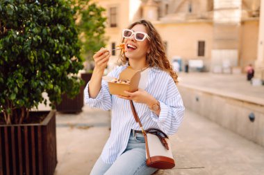 Neşeli bir kadın dışarıda aromatik makarna yer. Güneşli bir günde güzel bir turist bir paket kutusu tutar. Turizm, gıda ve eğlence kavramları.