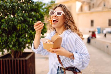Neşeli bir kadın dışarıda aromatik makarna yer. Güneşli bir günde güzel bir turist bir paket kutusu tutar. Turizm, gıda ve eğlence kavramları.