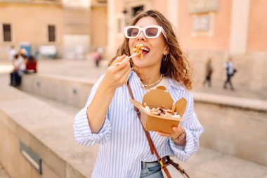 Neşeli bir kadın dışarıda aromatik makarna yer. Güneşli bir günde güzel bir turist bir paket kutusu tutar. Turizm, gıda ve eğlence kavramları.