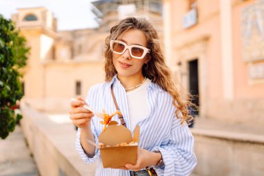 Neşeli bir kadın dışarıda aromatik makarna yer. Güneşli bir günde güzel bir turist bir paket kutusu tutar. Turizm, gıda ve eğlence kavramları.