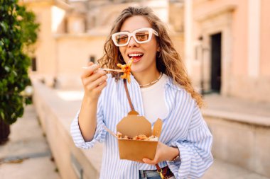 Neşeli bir kadın dışarıda aromatik makarna yer. Güneşli bir günde güzel bir turist bir paket kutusu tutar. Turizm, gıda ve eğlence kavramları.