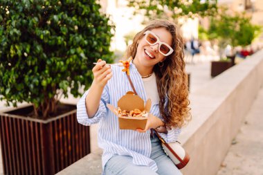 Neşeli bir kadın dışarıda aromatik makarna yer. Güneşli bir günde güzel bir turist bir paket kutusu tutar. Turizm, gıda ve eğlence kavramları.