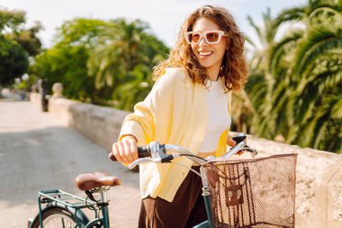 Şık giyinmiş neşeli bir kadın güneşli bir parkta bisiklet sürüyor. Güzel bir kadın bisiklet sürmekten hoşlanır. Turizm, eğlence, aktif yaşam tarzı kavramı.