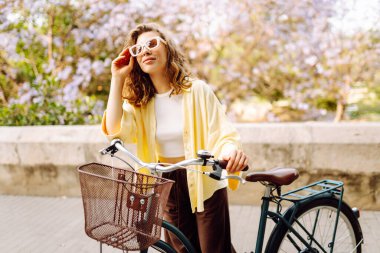 Şık giyinmiş neşeli bir kadın güneşli bir parkta bisiklet sürüyor. Güzel bir kadın bisiklet sürmekten hoşlanır. Turizm, eğlence, aktif yaşam tarzı kavramı.