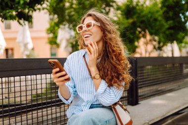 Güneş gözlüğü takan ve telefon kullanan gülümseyen kadın güneşli bir günde bankta oturur. Telefonunu kullanan genç bir kadın parkta yürümekten zevk alıyor ve bankta dinleniyor. İletişim ve rahatlama kavramları.