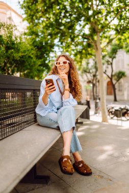 Güneş gözlüğü takan ve telefon kullanan gülümseyen kadın güneşli bir günde bankta oturur. Telefonunu kullanan genç bir kadın parkta yürümekten zevk alıyor ve bankta dinleniyor. İletişim ve rahatlama kavramları.