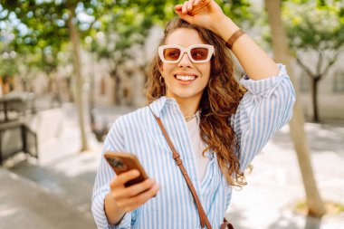 Güneş gözlüğü takan ve telefon kullanan gülümseyen kadın güneşli bir günde bankta oturur. Telefonunu kullanan genç bir kadın parkta yürümekten zevk alıyor ve bankta dinleniyor. İletişim ve rahatlama kavramları.